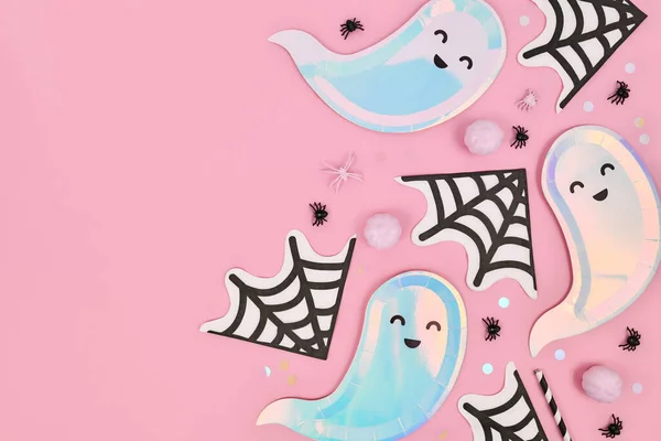 Cute pastel colored Halloween party flat lay with ghost shaped plates, spider web napkins and confetti on pink background