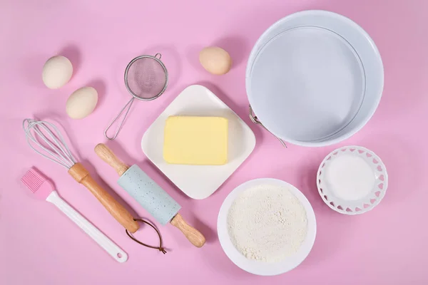 Cake dough ingredients and baking tools on pink background