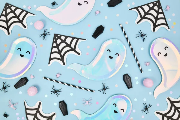 Cute pastel colored Halloween party flat lay with ghost shaped plates, spider web napkins and confetti on blue background