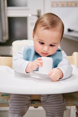 Bebek sandalyeye oturmuş yoğurt kapağıyla oynuyor.