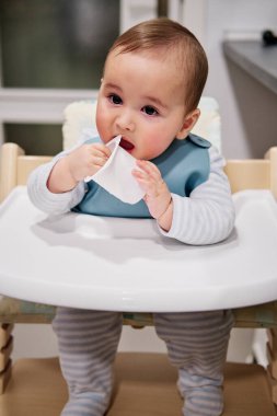 Bebek sandalyeye oturmuş yoğurt kapağıyla oynuyor.