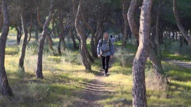 Man walking through the woods head-on. He wears a gray hat, a denim jacket, and a backpack on his back.