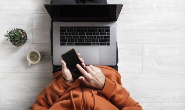 Evde çalış. Dizüstü bilgisayar ve akıllı telefon bir adamın elinde. İş adamı ya da serbest çalışan biri ahşap zeminde oturuyor..