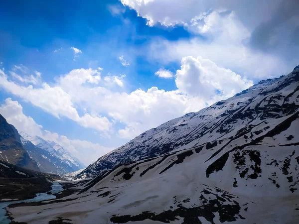 Karla kaplı dağlar, beyaz bulutlar ve mavi gökyüzü, kar dağının yanından akan nehir.