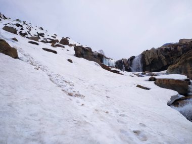 Mavi gökyüzünün önünde karla kaplı dağlar ve bir şelale. Karla kaplı dağ zirveleri. Kayalık kayaların üzerindeki bulutlar. Karda terk edilmiş dağ zirveleri. Resimli dağ manzarası.