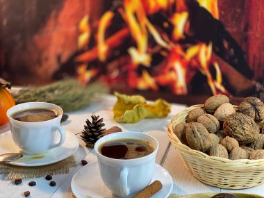 Beyaz ahşap bir masada sepetin içinde fındıklar var. Kahveli beyaz bir fincanın yanında. Uzaktan şöminenin alevlerini görebilirsiniz.