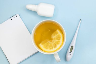 Elektronik termometrenin yanında mavi arka planda limon çayı ve burun spreyi olan beyaz bir bardak..