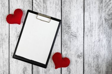 Ahşap gri zemin üzerinde beyaz boş bir çarşaf ve iki kırmızı kalp olan bir kırtasiye tableti. Mesaj için yer. Sevgililer Günü.