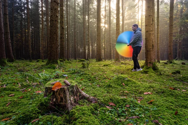 A lucky man is standing in the bright and mossy forest, turning his rainbow colored umbrella fast to have an colorful effect. Beautiful autumn landscapes with a heart leaf in the foreground.