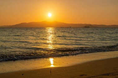 Turuncu güneş ışığı denize yansıyor, arka planda bir ada bulunan cennet gibi bir tatil teması.