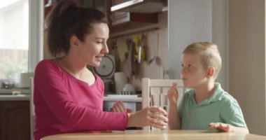 Çocuk, annesi mutfaktaki masada otururken bir hap alıyor ve su içiyor.