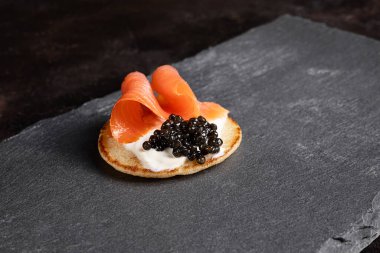 A small pancake with black sturgeon caviar and a slice of salmon on a slate