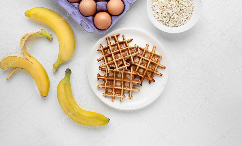 Sugar free waffles with banana and oatmeal, and ingredients for their preparation on a white table, healthy eating and gluten free breakfast concept, top view with copy space