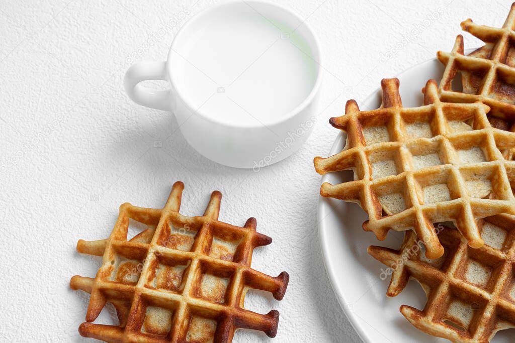 Freshly baked Belgian waffles on a white plate close-up and a cup of milk on a white table, tasty breakfast concept