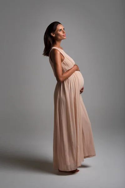Junge schöne schwangere Frau steht auf dem Boden trägt ein langes rosa Kleid vertikalen Blick grauen Hintergrund — Stockfoto