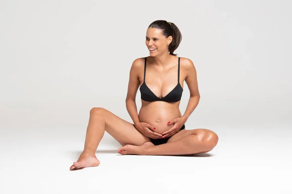 Junge schöne schwangere Frau sitzt auf dem Boden und zieht Unterwäsche an. horizontal weißer Hintergrund — Stockfoto