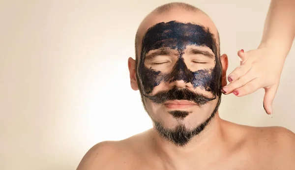 Man with closed eyes and black face mask. Woman puts exfoliating scrub on his face. Bald adult with beard and mustache. Concept health and wellness, skin care.
