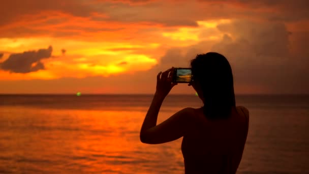 素晴らしいカラフルな夕日の空の写真を撮る若い魅力的な観光客の女の子.ビキニの女性ビーチ近くのスマートフォンで写真を撮る。高品質4Kスローモーショントロピカルライフ。タイのプーケット. — ストック動画
