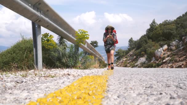 Ung Hiker Woman Använda Mobiltelefon Navigering eller sociala medier App. Flicka med ryggsäck går längs vägen och tittar på sin telefon. 4K Långsamma rörelser — Stockvideo