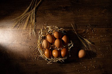 Bir sepet taze kahverengi yumurta ahşap bir zemin üzerinde dağınık buğday yan ışık ve tepeden aşağı fotoğraflanmış.