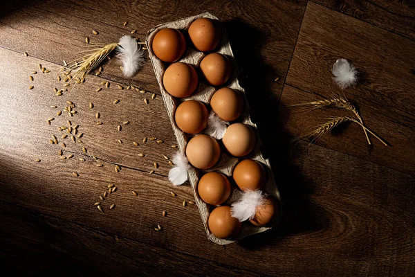 Bir düzine taze organik kahverengi yumurta dağınık tüy ve buğday ile yan ışık ve yukarıdan aşağı görüş ile dokulu bir arka plan karşı karşıya.