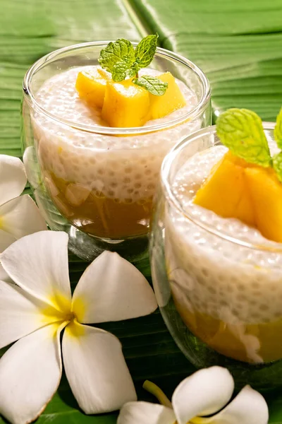 Tropical tapioca and coconut cream with mango dessert on green banana leaf background with frangipani flowers vertical format.