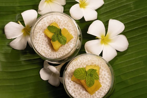 Tropical tapioca and coconut cream with mango dessert on green banana leaf background with frangipani flowers flat lay top down view.