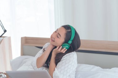 Genç Asyalı kadın yeşil kulaklık takıyor. Yatakta dizüstü bilgisayar kullanırken dinleniyor ve dinleniyor. Kız ses sesinden hoşlanıyor.