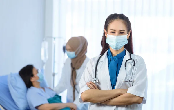 아시아인 의사는 얼굴에 마스크를 교차하여 환자를 배경으로 병원에서 일하는 — 스톡 사진