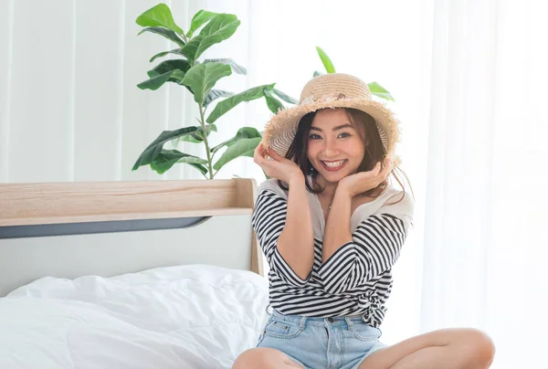 Portrait of smiling young Asian woman in summer casual clothes and straw hat sitting on bed at home. Cute asian girl