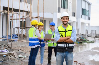 Şantiyede duran beyaz miğferli, yelekli, sakallı, genç ve çekici bir inşaat işçisinin portresi. Ev inşa kavramı