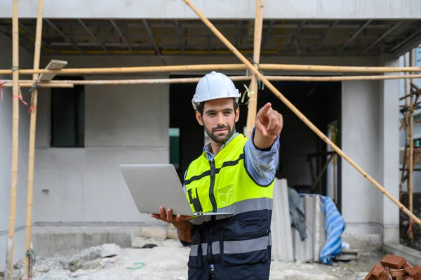 Young attractive construction smiling bearded in vest with white helmet working with laptop, standing on building construction site. Home building project