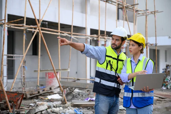 年轻迷人的男男女女身穿背心 头戴头盔 手持笔记本电脑 站在建筑工地上 住房建设项目 工程师工头在工作场所与同事讨论 — 图库照片