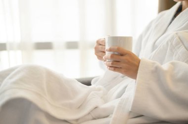 Bornozlu bir kadının beyaz bir fincan kahve içerken pencerenin yanında oturması ve işten ayrı olarak otelde harika bir sabah geçirmesi.