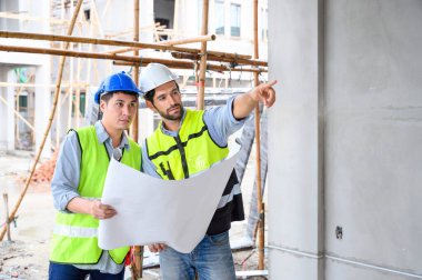 İnşaat mühendisi ve mimar kontrol planı inşaat alanının planıyla çalışıyor. Güvenlik için kasklı güvenlik yelekleri giyiyorlar. İnşaat projesi kavramı
