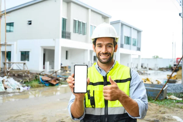 Young attractive construction man smiling bearded in vest with white helmet demonstrating blank smartphone and showing thumb up to the camera while standing on building construction site. White screen, mockup