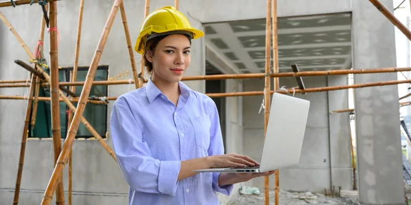 Young attractive construction woman smiling with yellow helmet working with laptop, standing on building construction site. Home building project