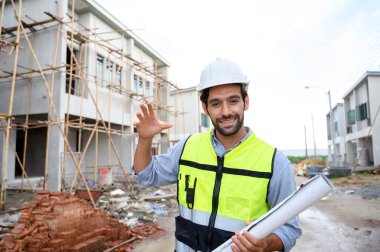 Young constructor engineer man smiling holding blueprint while talking about building construction, looking camera on under-construction building site. Happy Constructor bearded in vest with white helmet
