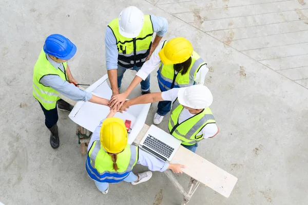 Top view of contractors, engineers and formats team in safety vests with helmets working with laptops, standing on under-construction building site. Home building project. Engineer foreman discusses with a coworker at workplace