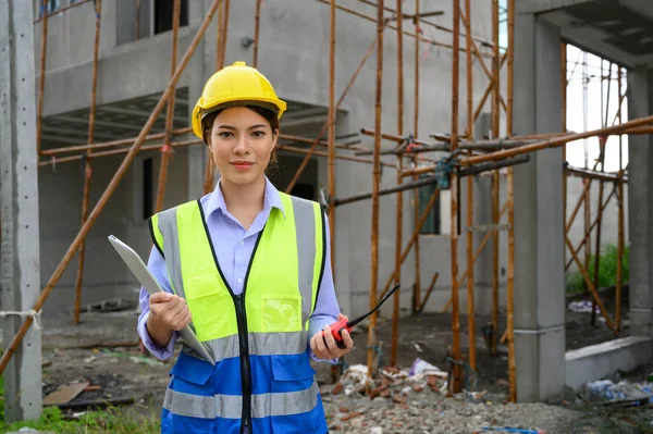 Young attractive construction woman smiling in vest with yellow helmet holding laptop and radio standing on building construction site. Home building project