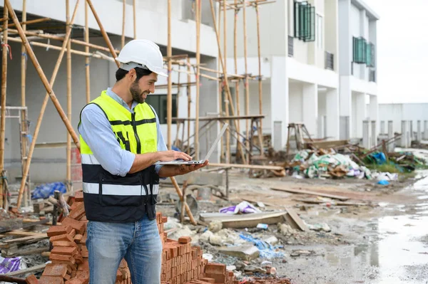 Young attractive construction smiling bearded in vest with white helmet working with laptop, standing on building construction site. Home building project