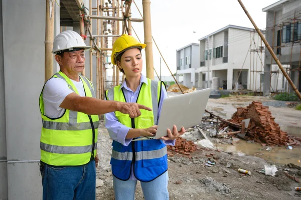 Construction woman and foreman in vest with helmet working with laptop, standing on under-construction building site. Home building project. Engineer foreman discusses with a coworker at workplace