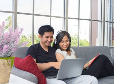 Genç Asyalı çift gündelik giysiler giyer oturma odasında rahat koltukta otururlar. Dizüstü bilgisayarda çalışan adam. Kulaklık takan ve tabletle oynayan bir kadın, ev konseptinde çalışıyor. WFH