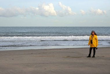 Kumsalda sarı yağmurluklu kadın. Sonbaharda rüzgarlı bir gün. Kuzey Denizi 'nin dalgaları kıyıya vuruyor