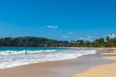 Sri Lanka. Mirissa Beach. Turquoise water and sea waves on a bright sunny day with a blue sky. People are swimming in the sea.