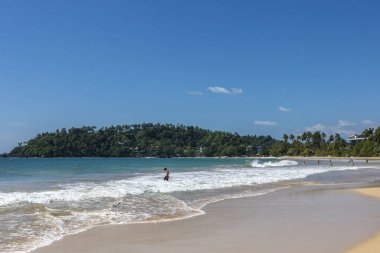 Sri Lanka. Mirissa Beach. Turquoise water and sea waves on a bright sunny day with a blue sky. People are swimming in the sea.
