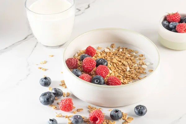 Bowl of granola with yogurt, milk and fresh raspberries, blueberries, mango for a healthy breakfast on a white marble table — Stock Photo, Image
