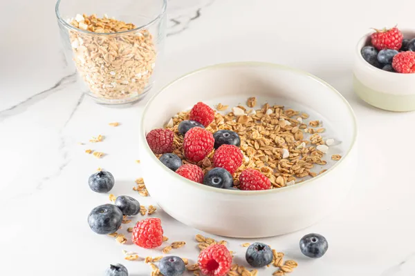 Egy tál granola joghurttal, tejjel és friss málnával, áfonyával, mangóval egy egészséges reggeliért egy fehér márványasztalon — Stock Fotó