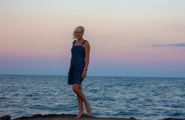 A beautiful blonde girl stands at sunset on the seashore of the ocean in a stylish denim dress, selective focus, travel vacation