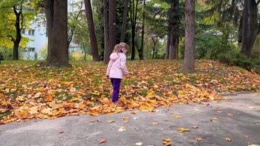 Şık kıyafetler içinde şirin küçük bir kız parkta yürüyor ve güneşli bir sonbahar gününde yaprakları topluyor, çocuklarla sağlıklı bir yaşam tarzı sürdürüyor.